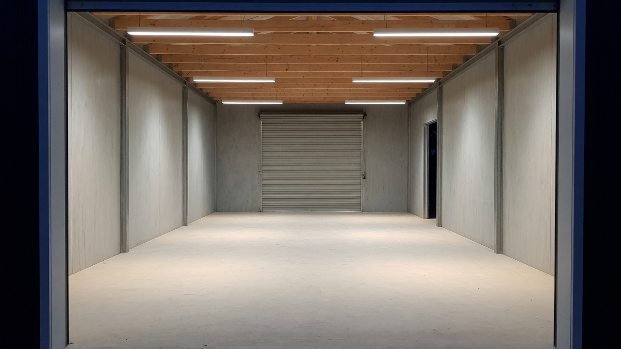 A generated placeholder image: an empty finished farm workshop at night, plywood lined, with six LED batten lights on overhead and the roller door closed at the far end.
