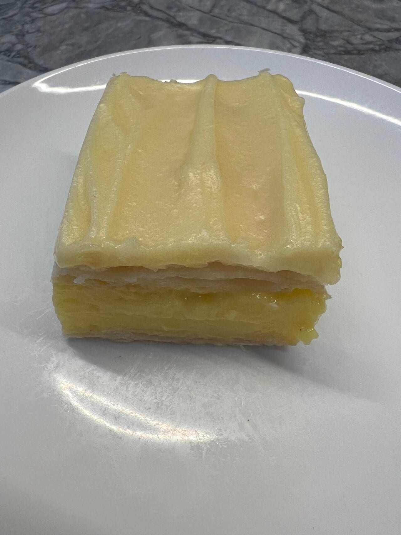 A custard square on a white plate: thick pale custard between two layers of flaky pastry, with a thin layer of icing on top.
