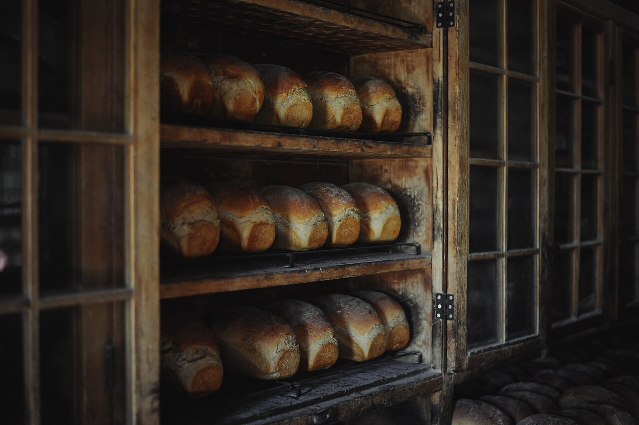 Rows of tin loaves with golden domed tops cooling on dark timber shelves behind glass doors, in low light.