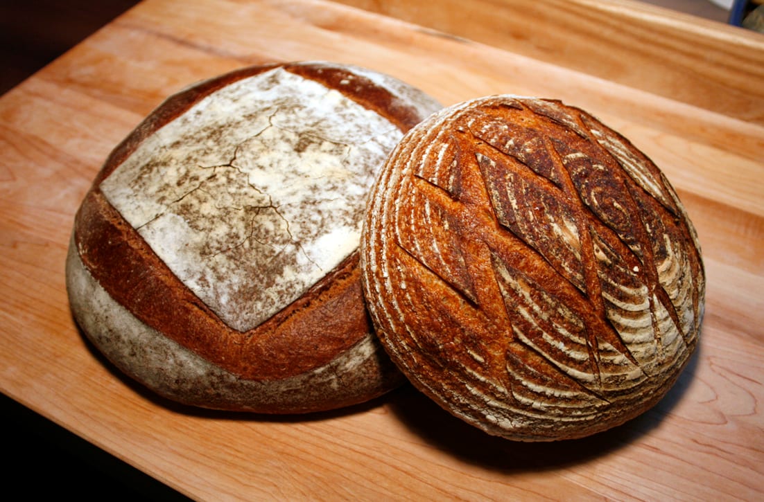 Two round sourdough loaves on a wooden board: one dusted with flour in a square pattern, the other scored in a diamond lattice with a dark crust.